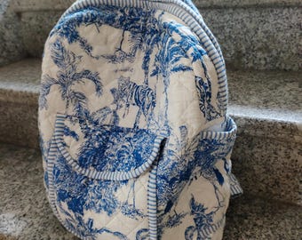 Peut inclure: Un sac à dos matelassé avec un motif de toile de Jouy bleu et blanc représentant une scène de jungle. Le sac à dos a une poignée supérieure, deux poches latérales et une bordure bleue rayée. Le motif principal comprend des tigres et des palmiers.