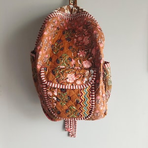 May include: A quilted backpack in shades of brown, adorned with a floral pattern in pink, green, and blue. The bag features a front pocket, side pockets, and a decorative tassel. White and red striped trim accents the edges.
