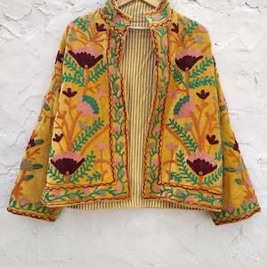 May include: Mustard yellow embroidered jacket with floral patterns in pink, green, and burgundy. The jacket has a striped lining and is hanging on a wooden hanger against a white textured wall. The embroidery details are intricate.