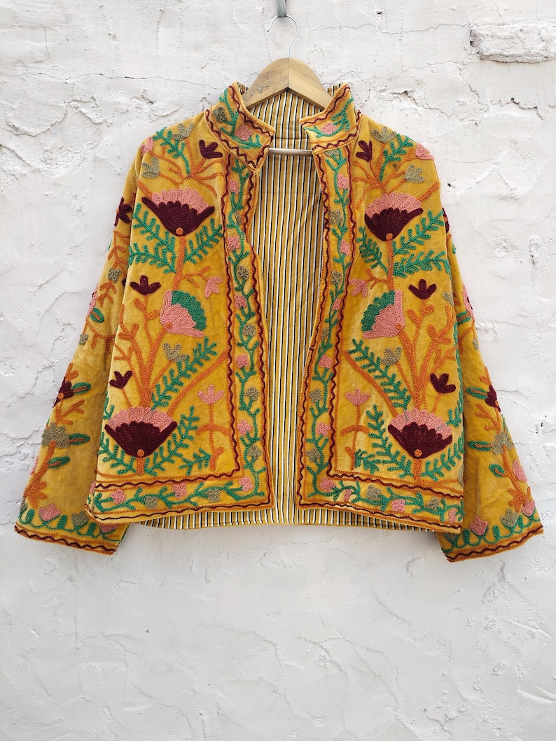 May include: Mustard yellow embroidered jacket with floral patterns in pink, green, and burgundy. The jacket has a striped lining and is hanging on a wooden hanger against a white textured wall. The embroidery details are intricate.