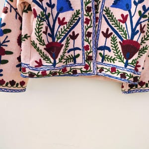 May include: A pink embroidered jacket with blue, green, and red floral patterns. The jacket has a button closure and long sleeves.