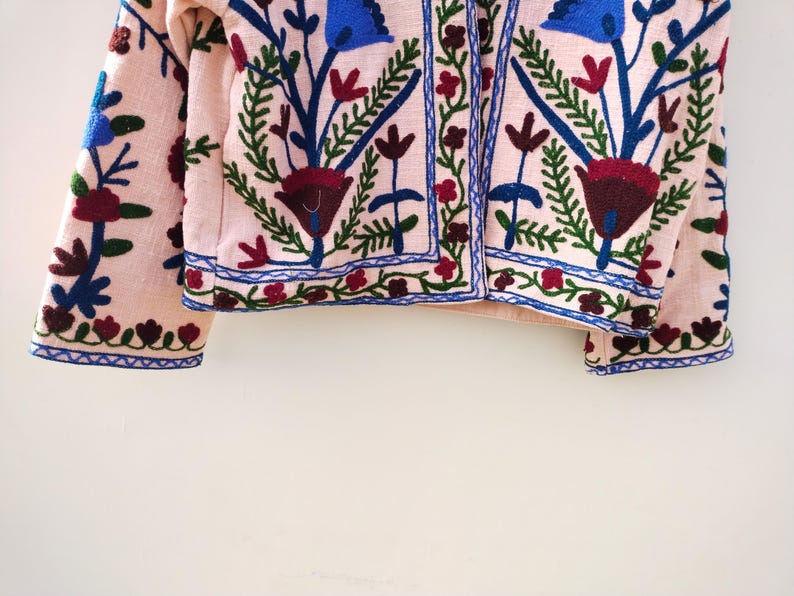 May include: A pink embroidered jacket with blue, green, and red floral patterns. The jacket has a button closure and long sleeves.