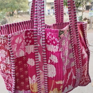 May include: A patchwork tote bag in shades of pink and red, featuring floral and geometric patterns. The bag has sturdy handles with vertical stripes and a rectangular shape, perfect for carrying items.