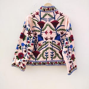 May include: A pink embroidered jacket with a floral pattern in shades of blue, red, and green. The jacket has a mandarin collar and long sleeves.