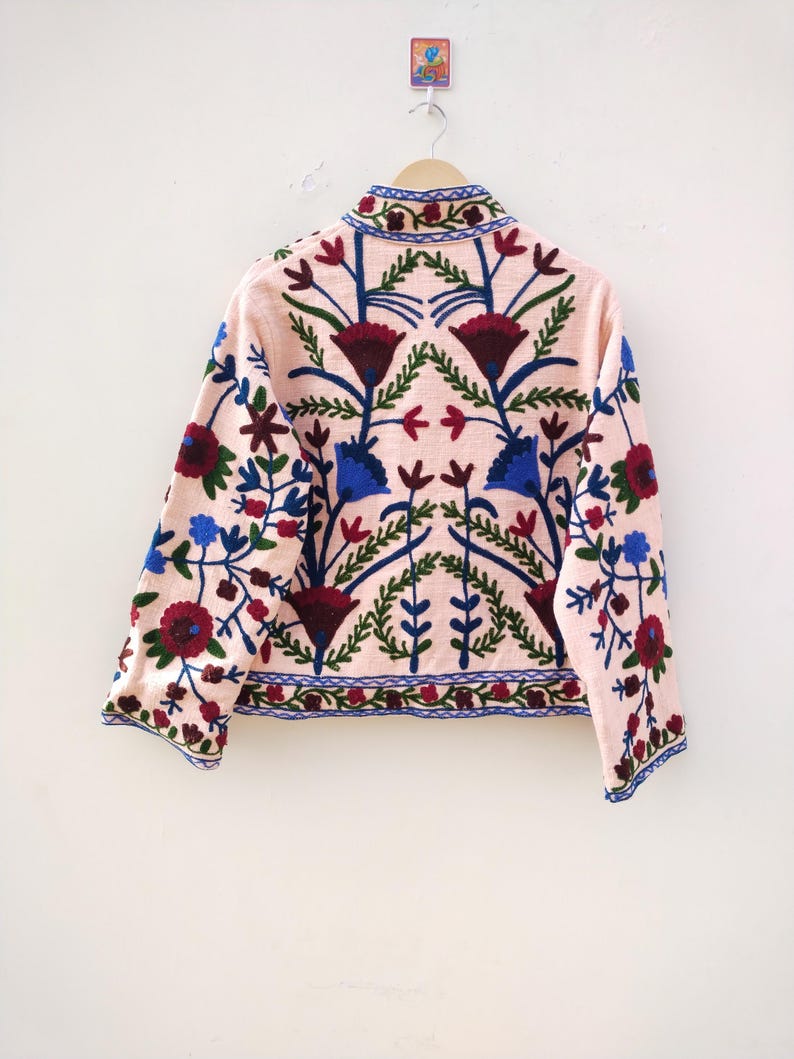 May include: A pink embroidered jacket with a floral pattern in shades of blue, red, and green. The jacket has a mandarin collar and long sleeves.