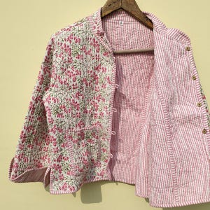 May include: A quilted jacket with a floral pattern on the exterior and a striped interior. The jacket is mainly white with pink floral designs and a pink and white striped lining. It has a button-up front and two pockets.