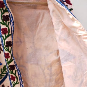 May include: A cream-colored jacket with a blue, green, and red floral embroidery design. The jacket is lined with a cream-colored fabric.