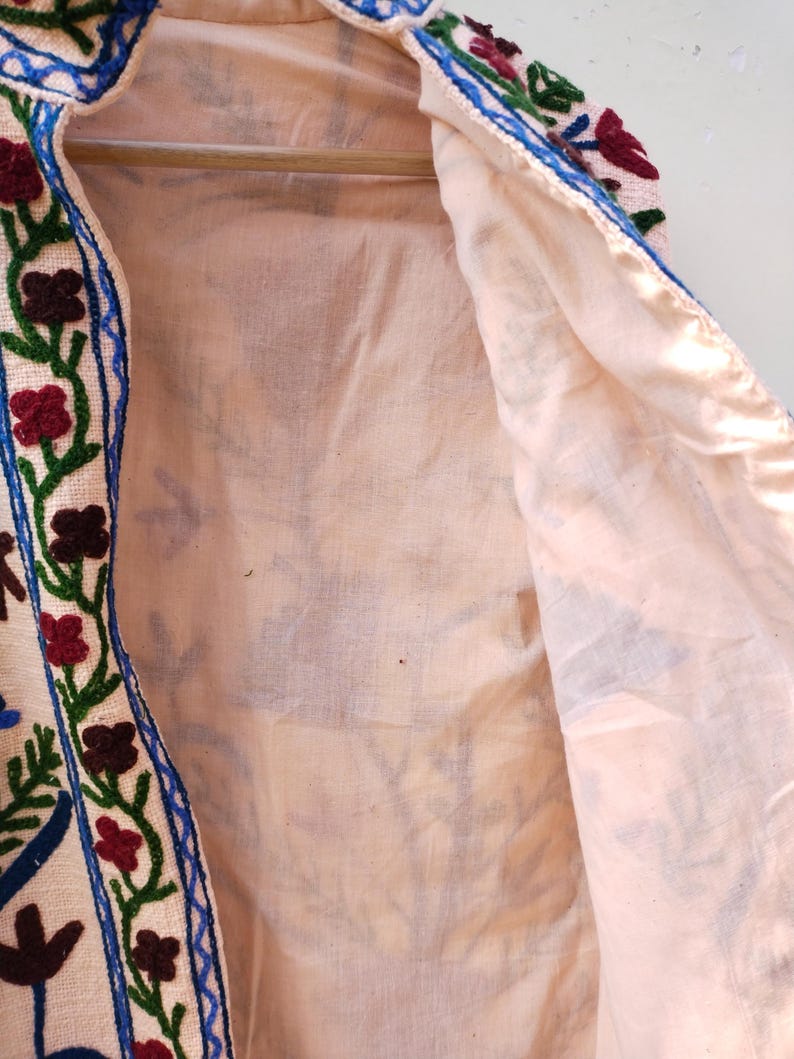 May include: A cream-colored jacket with a blue, green, and red floral embroidery design. The jacket is lined with a cream-colored fabric.