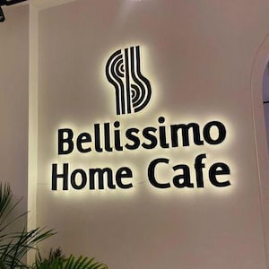 May include: A wall-mounted sign for "Bellissimo Home Cafe" with a black logo and text, illuminated against a white wall. A green plant is in the foreground, and a light fixture hangs from the ceiling.