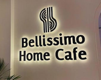 May include: A wall-mounted sign for "Bellissimo Home Cafe" with a black logo and text, illuminated against a white wall. A green plant is in the foreground, and a light fixture hangs from the ceiling.