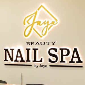 May include: Interior shot of a nail spa with a sign that reads "Jaya Beauty Nail Spa". The sign is illuminated with gold and white lettering. The spa has wooden accents and shelves displaying nail polish colors.