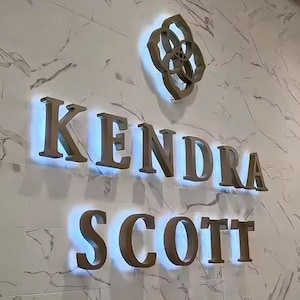 May include: A wall-mounted sign featuring the Kendra Scott logo and brand name in metallic gold lettering. The letters and logo are backlit with a bright white glow, mounted against a white wall with a grey marble-like veined pattern throughout.
