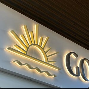 May include: A backlit wall sign featuring a stylized sun and waves design in yellow and white. The word "GO" is also visible, with a similar illuminated effect. The sign is mounted on a white wall.