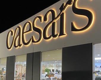 May include: A building exterior featuring a large, black, three-dimensional sign that reads "caesar's" in a cursive font. The letters are mounted on a white wall and are backlit with a warm, glowing light that creates a halo effect around each character.