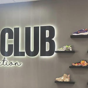 May include: A gray wall features the large black backlit text "THE CLUB" above the word "Collection" in a cursive font. Six individual black floating shelves are mounted to the right, each displaying a different athletic sneaker in various colors.