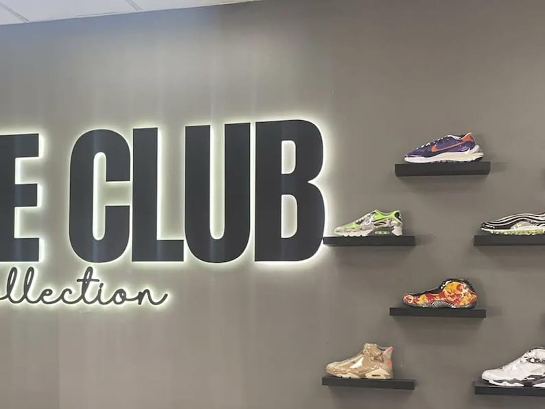 May include: A gray wall features the large black backlit text "THE CLUB" above the word "Collection" in a cursive font. Six individual black floating shelves are mounted to the right, each displaying a different athletic sneaker in various colors.
