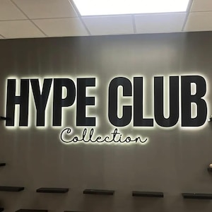 May include: A wall display featuring the text HYPE CLUB Collection in large, bold, black letters with a white backlit glow. The display is mounted on a dark gray wall, flanked by several small, black, horizontal shelves holding various sneakers.