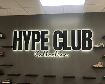 May include: A wall sign for "HYPE CLUB Collection" with black block letters and a glowing white outline. The word "Collection" is in a cursive font. The sign is above shelves displaying sneakers.