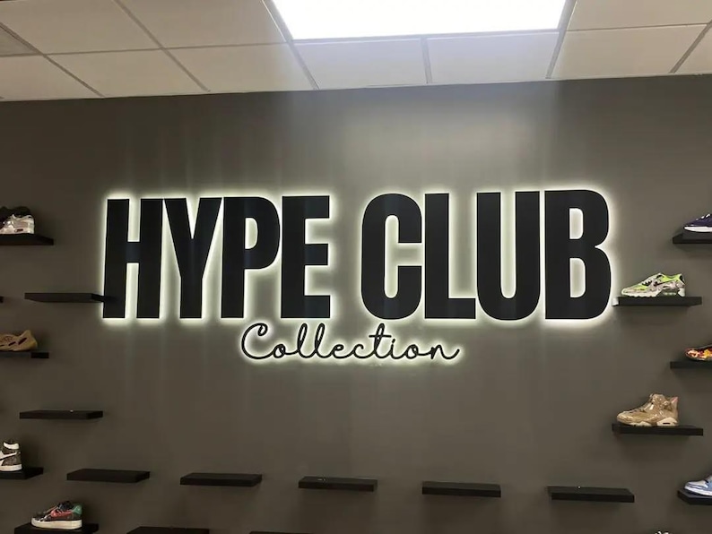 May include: A wall display featuring the text HYPE CLUB Collection in large, bold, black letters with a white backlit glow. The display is mounted on a dark gray wall, flanked by several small, black, horizontal shelves holding various sneakers.