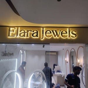 May include: A storefront sign featuring the gold-colored, backlit 3D lettering 'Elara Jewels' mounted on a horizontal panel. The letters are illuminated from behind, creating a glowing effect against the wall above the glass entrance.