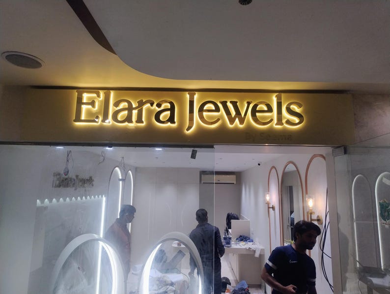 May include: A storefront sign featuring the gold-colored, backlit 3D lettering 'Elara Jewels' mounted on a horizontal panel. The letters are illuminated from behind, creating a glowing effect against the wall above the glass entrance.
