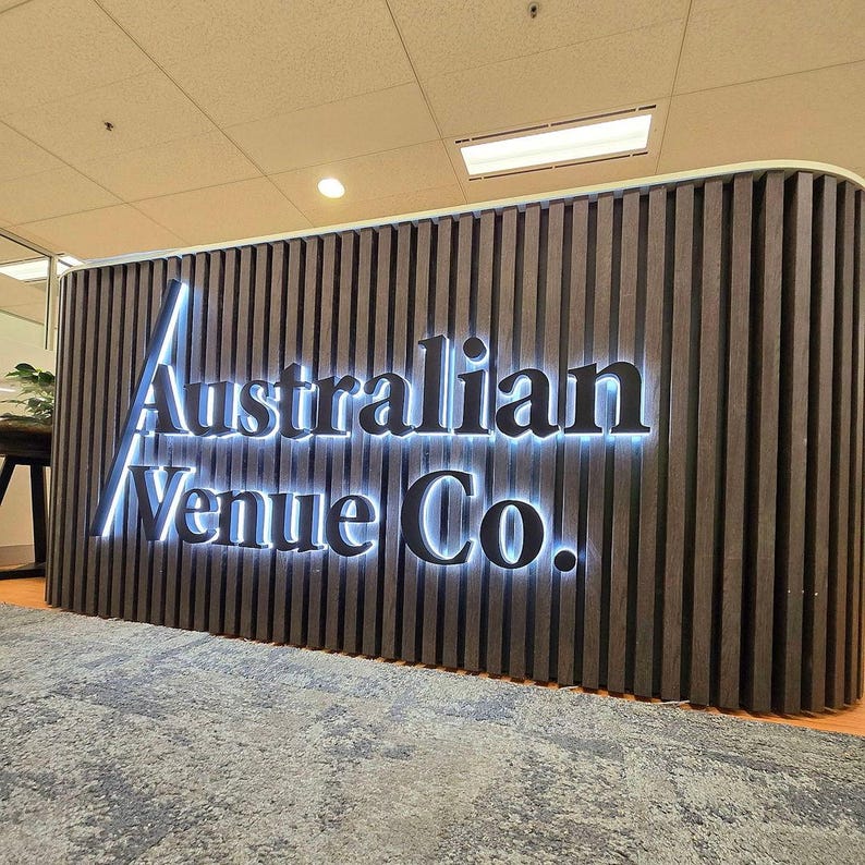 May include: A curved reception desk featuring dark brown vertical wooden slats and a large, backlit sign that reads Australian Venue Co. in black, dimensional lettering. The sign is mounted directly onto the slatted wooden surface.
