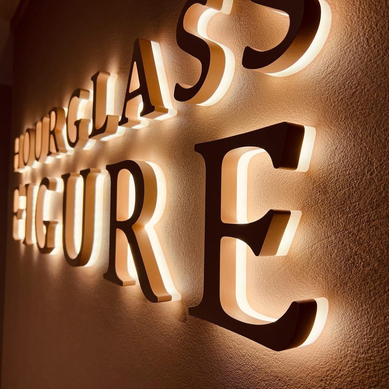 May include: A wall-mounted sign featuring the words HOURGLASS FIGURE in large, dark, three-dimensional capital letters. Each letter is backlit with a warm white glow, creating a halo effect against the textured surface of the wall.