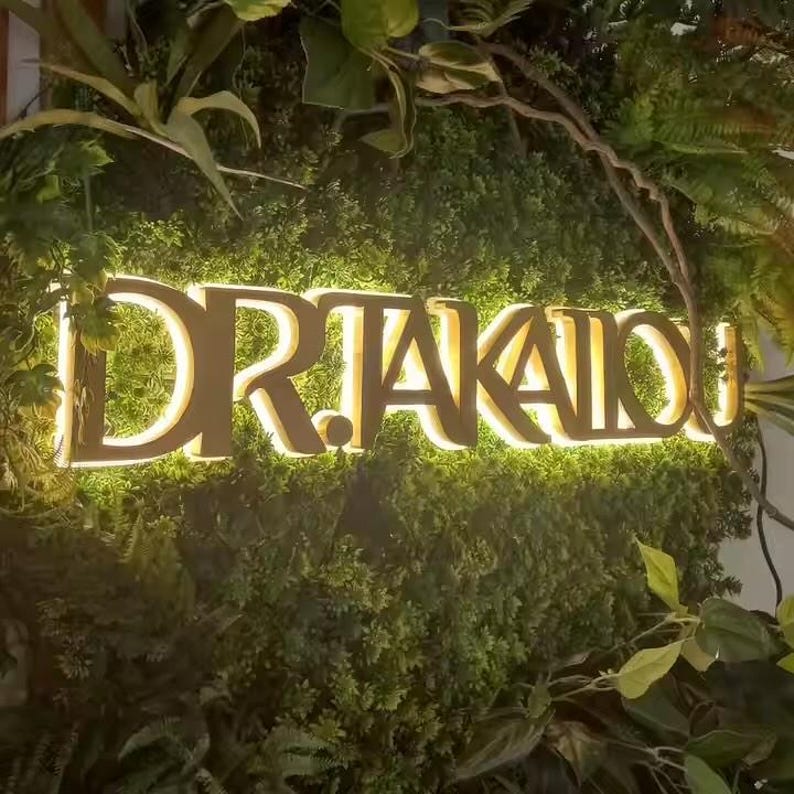 May include: A three-dimensional sign featuring the text DR.TAKAIOU in bold, gold-toned capital letters. The sign is backlit with a warm glow and is mounted against a dense, textured wall covered in various shades of green artificial foliage.