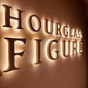 May include: A wall-mounted sign featuring the words HOURGLASS FIGURE in large, dark, three-dimensional capital letters. Each letter is backlit with a warm, glowing light that creates a soft halo effect against the textured wall surface behind it.