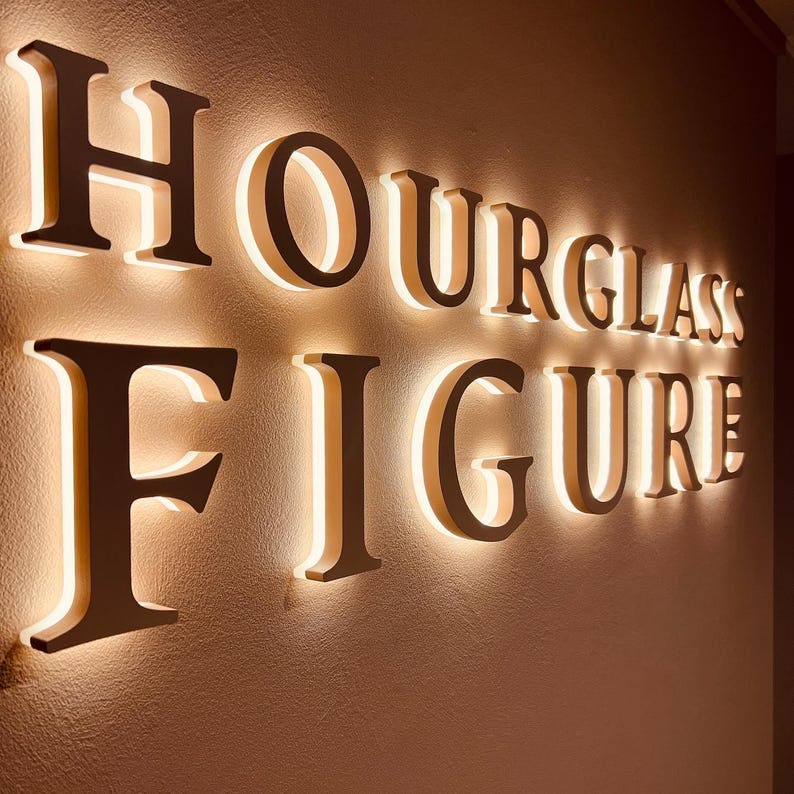 May include: A wall-mounted sign featuring the words HOURGLASS FIGURE in large, dark, three-dimensional capital letters. Each letter is backlit with a warm, glowing light that creates a soft halo effect against the textured wall surface behind it.