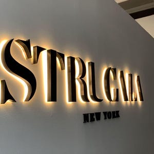 May include: A three-dimensional wall sign featuring the black lettering STRUGALA with a warm, glowing backlight effect. Below the main text, the words NEW YORK are displayed in smaller, non-illuminated black letters against a light wall.