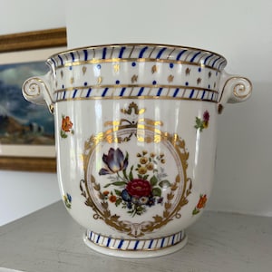 May include: White ceramic vase with gold trim and floral designs. The vase features a central floral medallion and small floral accents. Blue and gold bands encircle the top and base, with two curved handles.