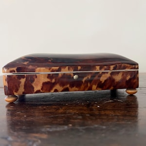 May include: A rectangular box with a glossy, mottled brown and tan tortoiseshell pattern. The box features a smooth, curved lid, a thin silver-toned metal trim along the center seam, and four small, rounded, light-colored feet at the base.