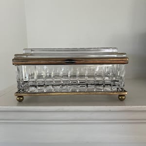 May include: A rectangular glass box with a clear, faceted geometric pattern on the sides. The container features a metallic bronze-toned frame and a hinged lid, supported by four small, rounded feet in a matching bronze finish.