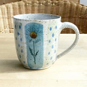 Könnte beinhalten: Eine gesprenkelte Keramiktasse mit großem Henkel. Die Tasse zeigt ein gemaltes Gänseblümchen-Design auf einem hellblauen vertikalen Streifen, umgeben von blauen Tupfen. Die Tasse steht auf einer hellen Holzoberfläche.