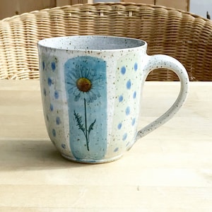 Könnte beinhalten: Eine weiße Keramiktasse mit einem blauen und grünen Gänseblümchen-Design. Die Tasse hat eine gesprenkelte Glasur und einen hellblauen Henkel.