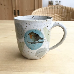 Könnte beinhalten: Eine weiße Keramiktasse mit braunen Flecken und einem blauen Kreis mit einem Rotkehlchen, das auf einem Ast sitzt. Die Tasse hat einen Henkel und ist mit einem Muster aus hellgrünen und grauen Kreisen verziert.