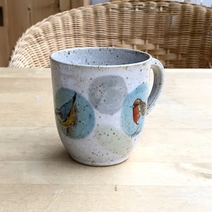 Könnte beinhalten: Eine gesprenkelte Keramiktasse mit Henkel, die Illustrationen von Vögeln in blauen und grauen Kreisen zeigt. Die Tasse hat eine hellgrau gesprenkelte Außenseite und steht auf einer Holzoberfläche.