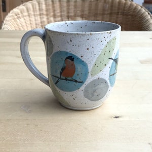 Könnte beinhalten: Eine weiße Keramiktasse mit gesprenkeltem Überzug zeigt ein abstraktes Design in Blau, Grün und Grau mit zwei Vögeln. Ein Vogel ist orange und schwarz, der andere grau und schwarz.