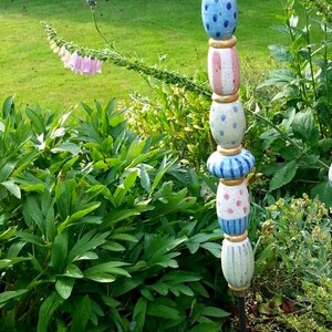 May include: A colorful, handcrafted garden stake with a blue teardrop-shaped top. The stake features stacked ceramic beads in blue, pink, and white with dot and stripe patterns. The stake is approximately 60 inches tall.