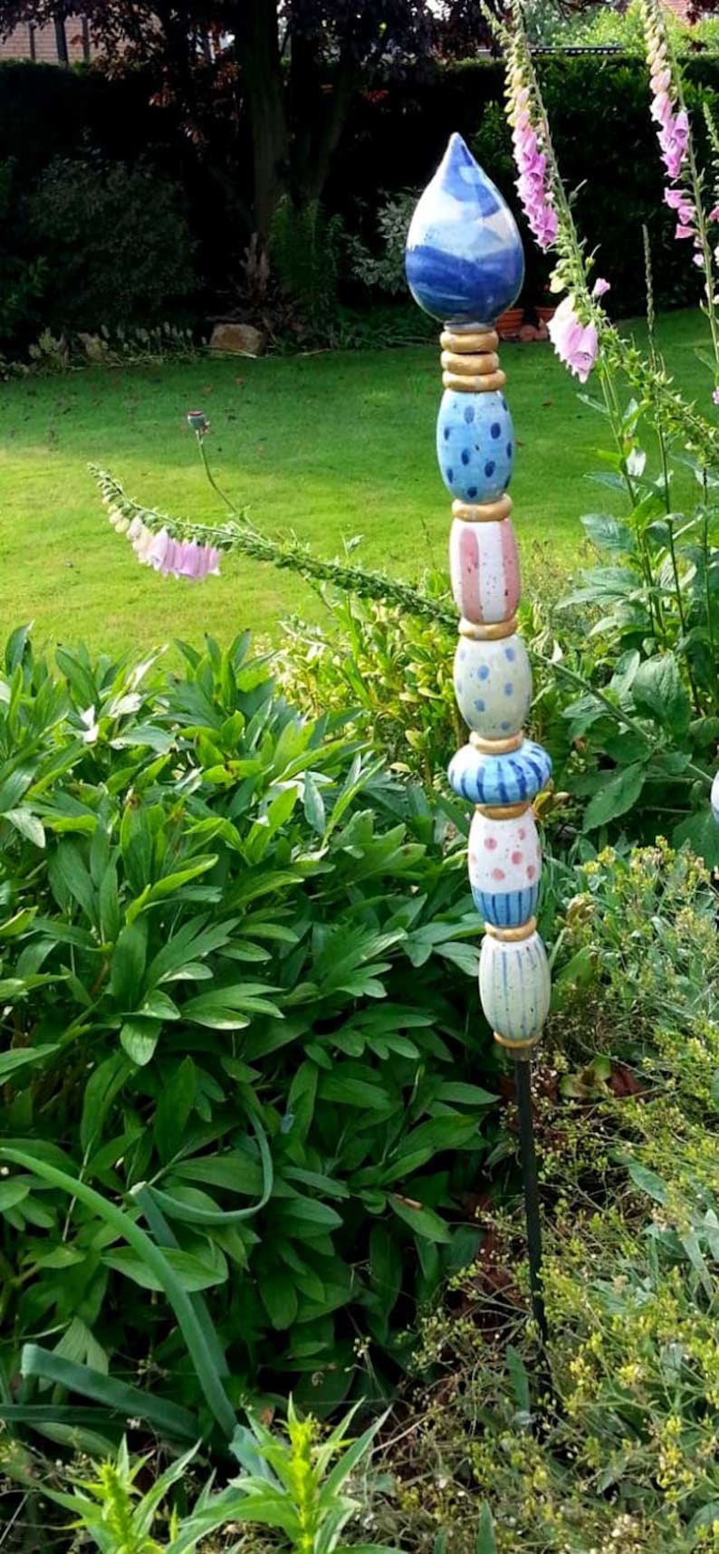 May include: A colorful, handcrafted garden stake with a blue teardrop-shaped top. The stake features stacked ceramic beads in blue, pink, and white with dot and stripe patterns. The stake is approximately 60 inches tall.