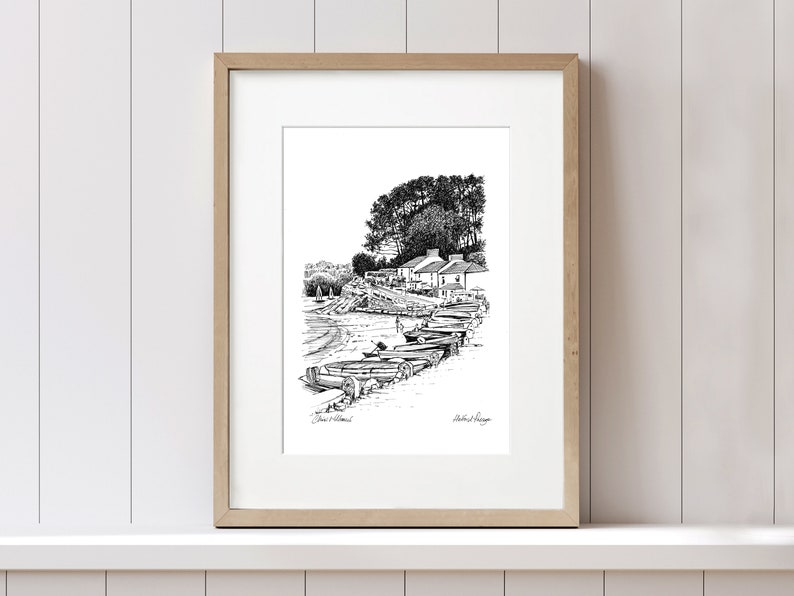 Puede incluir: Dibujo en tinta negra y blanca de un peque&ntilde;o pueblo con una fila de botes en la orilla. El dibujo est&aacute; enmarcado en un marco de madera claro.