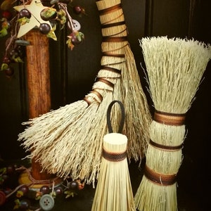 May include: Three handmade brooms with natural straw bristles and brown and black twine accents. The brooms are displayed against a dark wood background.