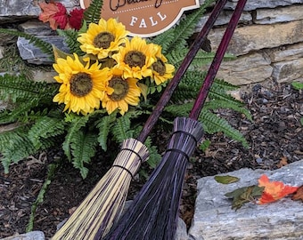 May include: Two brooms with black and white bristles and wooden handles. The brooms are leaning against a stone wall with a sign that says "Hello Beautiful Fall".