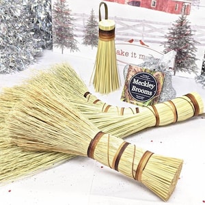 May include: Several natural fiber brooms with brown and tan accents. One broom has a loop handle. The brooms are displayed with a holiday-themed backdrop featuring a red barn and snow-covered trees. A small package of "Meckley Brooms" is also visible.