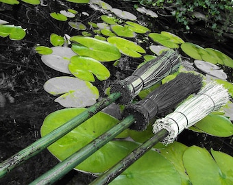May include: Three brooms with different colored bristles are lying on top of green lily pads in a pond. The brooms are made of natural materials and have wooden handles.