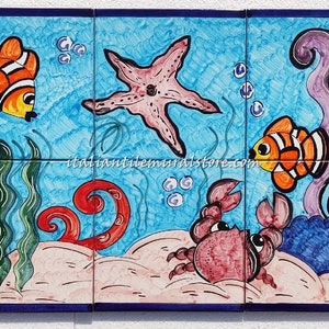 Op de afbeelding: Een kleurrijke keramische tegelmuur die een onderwater scène afbeeldt met rode en roze octopussen, oranje en witte clownvissen, een roze zeester, een rode krab en groene en paarse zeewier. De tegelmuur is tegen een blauwe achtergrond geplaatst.