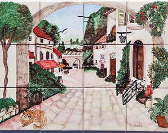 May include: A hand-painted ceramic tile mural depicting a quaint Italian village scene. The mural features a white building with a brown door, a red-roofed building, and a lush green courtyard with a cat sitting on the ground. The tile mural is perfect for adding a touch of European charm to any home.