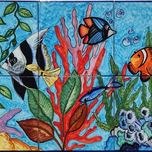 Op de afbeelding: Een kleurrijke keramische tegelmuur die een onderwater scène voorstelt met verschillende vissen, koraal en zeewier. De muurschildering toont een zwart-witte engelvis, een blauwe vis, een clownvis en rood koraal. De tegelmuur is gesigneerd met "Kaléidos"