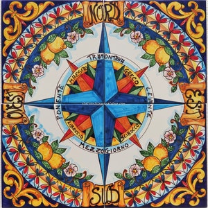 May include: A colourful Italian ceramic tile featuring a compass design. The compass points to the four cardinal directions: North, South, East, and West. The tile is decorated with lemons, flowers, and scrolls.